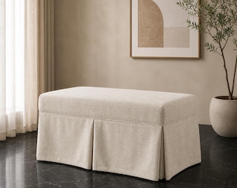 Pode incluir: Um otomano retangular estofado em tecido bege claro, com uma superfície superior lisa e uma base com saia plissada. A peça apresenta um design estruturado com linhas limpas e uma paleta de cores neutras, sendo um item de mobiliário funcional.