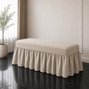 May include: A rectangular bench upholstered in a solid light beige fabric. The bench features a flat, cushioned top and a gathered, floor-length skirt that drapes around the base, creating a uniform and structured appearance in a neutral tone.