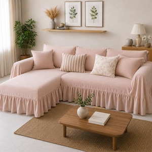 Op de afbeelding: Een lichtroze hoekbank met een gefranjerde rok. De bank is versierd met verschillende sierkussens in verschillende patronen en roze tinten. Een kleine houten salontafel en een geweven vloerkleed staan voor de bank.