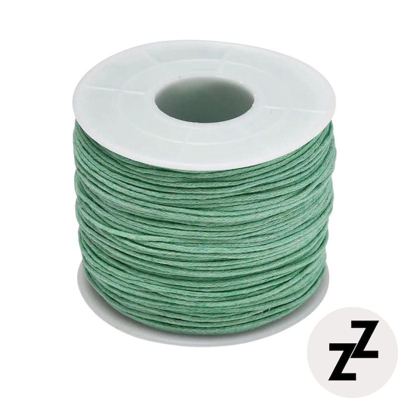 May include: A spool of green waxed cord. The cord is tightly wound around a white plastic spool. The cord is a muted green color. The spool has a central hole and a circular base. The image is on a white background.
