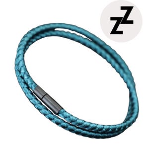 Puede incluir: Pulsera trenzada de color turquesa con un cierre gris oscuro. La pulsera es circular y presenta un diseño de doble hebra. Un círculo blanco con la letra negra "Z" repetida dos veces está en la esquina superior derecha.
