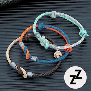 Peut inclure: Trois bracelets en corde colorés avec des détails noués. Les bracelets présentent des nuances de bleu, d'orange, de marron et de gris. Un bracelet a un petit charme doré. Les bracelets sont présentés sur une surface sombre et texturée.