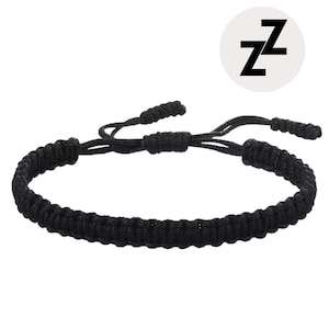 Puede incluir: Pulsera trenzada negra con nudos corredizos ajustables. La pulsera está hecha de un material grueso y tejido. Un círculo blanco con la letra "Z" en negro está en la esquina superior derecha.