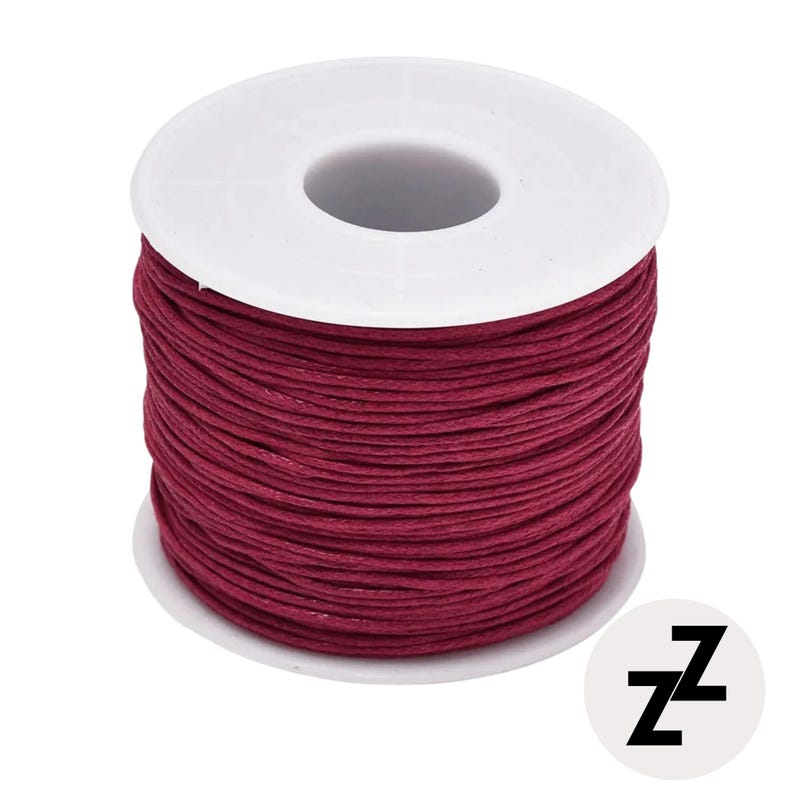 May include: A spool of burgundy-colored thread. The thread is tightly wound around a white plastic spool. A small white circle with the black letters "zz" is in the lower right corner.