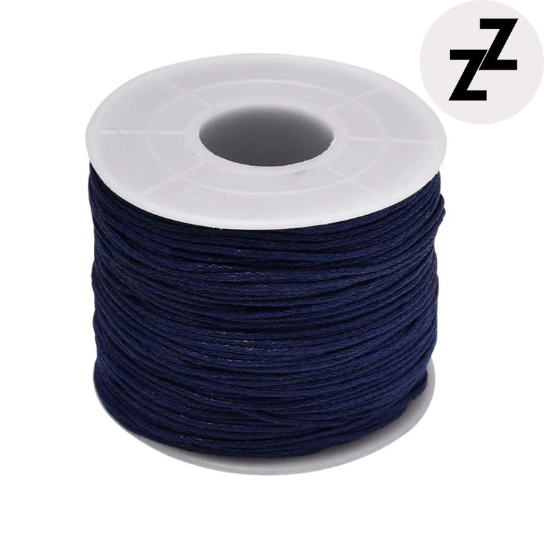 May include: A spool of dark blue waxed cord. The cord is tightly wound around a white plastic spool. The spool has a central hole and a circular design. A white circle with the letter "Z" is in the upper right corner.