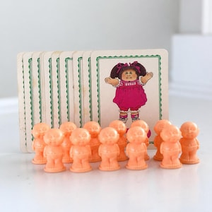 May include: A set of small, peach-colored plastic figurines arranged in a row in front of a stack of rectangular playing cards. Each card features a printed illustration of a cartoon character wearing a pink outfit with dark brown hair.