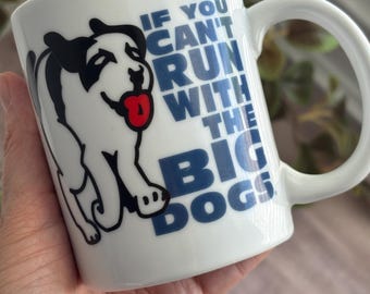 May include: A white ceramic mug featuring a black and white graphic of a dog with a red tongue. The text printed on the mug reads, IF YOU CAN'T RUN WITH THE BIG DOGS, in bold blue capital letters. The mug has a curved handle on the right side.