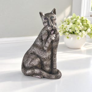 May include: A silver-toned cat figurine in a seated position, grooming its paw with a small pink tongue. The sculpture features a textured, weathered finish with dark recessed details and prominent, almond-shaped purple eyes.