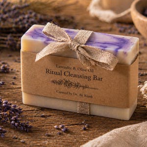 May include: A rectangular bar of soap with a lavender and olive oil blend, wrapped in a brown paper band and tied with a ribbon. The label reads "Ritual Cleansing Bar". Dried lavender and a wooden bowl are in the background.