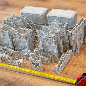 Peut inclure: Un ensemble de ruines de bâtiments miniatures et de sections de murs imprimés en 3D gris clair avec des détails architecturaux complexes, disposés à côté d'un mètre ruban jaune indiquant une longueur d'environ 43 centimètres.