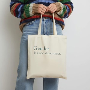 May include: Cream-colored tote bag with the text "Gender is a social construct" in dark green. The bag has two handles and is held by a person wearing a striped sweater and blue jeans.