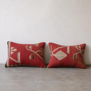Könnte beinhalten: Zwei rechteckige Dekokissen mit einer tiefroten Grundfarbe und eingewebten geometrischen Mustern in Beige und Braun. Der strukturierte Stoff zeigt kantige Formen und Linien auf der Vorderseite beider Kissen, die identisch gestaltet sind.