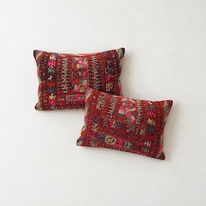 May include: Two rectangular throw pillows featuring a deep red base colour with intricate, multi-coloured embroidery. The patterns consist of geometric shapes, floral motifs, and linear stitching in shades of pink, yellow, blue, and white.