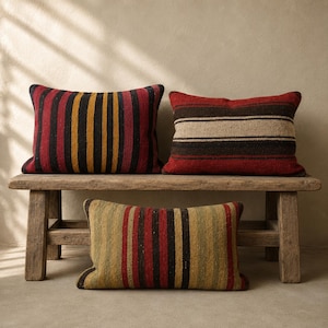 May include: Three rectangular throw pillows featuring woven striped patterns. Two pillows sit on a wooden bench, one with vertical red, black, and yellow stripes, the other with horizontal red, brown, and beige stripes. A third pillow with vertical red, black, and olive green stripes rests below.