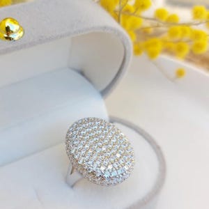 Puede incluir: Un anillo de plata con una gran cara ovalada cubierta de pequeñas piedras claras y brillantes. El anillo se muestra en una caja de joyería de terciopelo gris, con flores amarillas en el fondo.