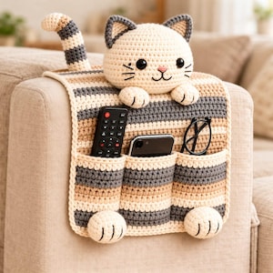 Può includere: Organizzatore per bracciolo del divano a forma di gatto all'uncinetto. L'organizzatore a righe beige, grigie e marroni ha tre tasche che contengono un telecomando, un telefono e occhiali. Il gatto ha un viso sorridente, orecchie e zampe.