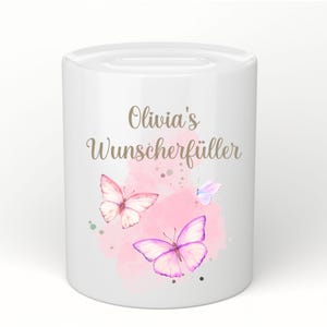 May include: White ceramic money bank with a slot on top. The bank is decorated with watercolor butterflies in pink and purple hues, and the text "Olivia's Wunscherf&uuml;ller" in gold script.