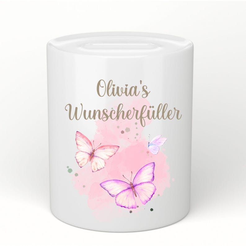 May include: White ceramic money bank with a slot on top. The bank is decorated with watercolor butterflies in pink and purple hues, and the text "Olivia's Wunscherf&uuml;ller" in gold script.