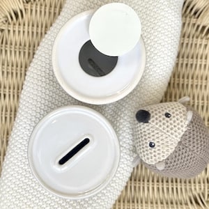 May include: Overhead shot of two white ceramic piggy bank lids with coin slots, one with a white disc on top. A small, knitted hedgehog toy with a gray and beige body and black eyes rests nearby. The items are on a white knitted blanket.