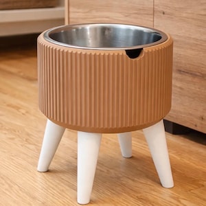 Könnte beinhalten: Ein moderner Futternapf für Haustiere mit einem Edelstahl-Innenraum und einer hellbraunen, geriffelten Außenseite. Die Schüssel steht auf drei weißen, abgewinkelten Beinen. Das Design ist schlicht und stilvoll, passend für jede Einrichtung.