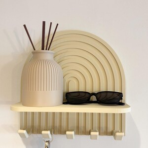 Peut inclure: Une étagère murale beige dotée d'un panneau arrière arqué avec des crêtes semi-circulaires concentriques. L'étagère comprend cinq crochets sur le bord inférieur, un vase côtelé contenant des bâtonnets marron foncé et des lunettes de soleil noires.