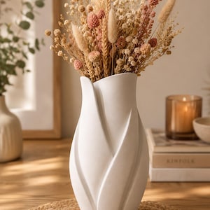 Op de afbeelding: Een witte keramische vaas met een sculpturaal, overlappend bloembladontwerp, gevuld met een boeket gedroogde bloemen in tinten beige, zand en gedempt roze. De vaas heeft een gladde, matte afwerking en een taps toelopend silhouet dat naar het midden toe uitloopt.