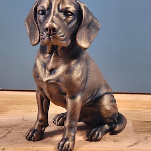 Könnte beinhalten: Bronze-farbene Hundestatue in sitzender Position. Die Skulptur weist detaillierte Fell- und Gesichtszüge auf, einschließlich Schlappohren und einem fokussierten Blick. Die Pfoten des Hundes sind sichtbar, und die Statue hat ein metallisches Finish.