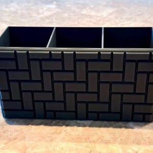 Peut inclure: Un organiseur de bureau rectangulaire gris foncé avec trois compartiments. L'extérieur présente un motif en forme de briques. L'organisateur est conçu pour contenir des stylos, des crayons ou d'autres fournitures de bureau. L'article est fait d'un matériau solide.