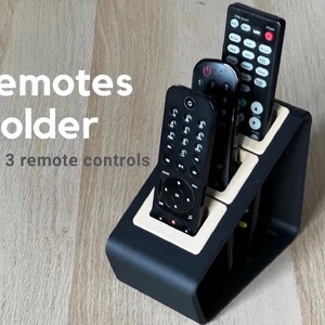 Può includere: Porta telecomandi nero con tre telecomandi neri. Il supporto ha un bordo beige e il testo "remotes holder for 3 remote controls". I telecomandi hanno vari pulsanti.