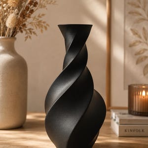 Peut inclure: Un vase noir mat doté d'une silhouette torsadée en spirale. Le vase présente un col étroit qui s'élargit en un corps arrondi et profilé, avec des rainures profondes et incurvées créant un effet visuel dynamique et rotatif sur la surface.