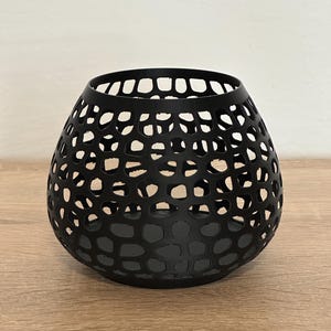 Peut inclure: Un bougeoir noir mat présentant une forme arrondie et bulbeuse avec un motif en treillis ajouré complexe rappelant des structures cellulaires organiques. Le récipient possède une large ouverture circulaire au sommet et une base pleine.