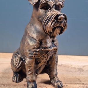 Pode incluir: Estátua de cão Schnauzer na cor bronze, em posição sentada. A estátua apresenta detalhes, incluindo um pelo texturizado, uma longa barba e orelhas caídas. O cão está sobre uma superfície de madeira com um fundo azul-acinzentado.