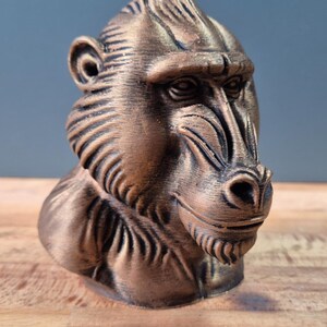 May include: A bronze-colored sculpture of a mandrill's head and upper torso. The sculpture has detailed features, including the mandrill's distinctive facial ridges and prominent nose. The sculpture is displayed on a wooden surface.