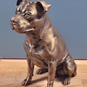 Peut inclure: Statue de chien de couleur bronze, probablement un Staffordshire Bull Terrier, assis. Le chien a une musculature prononcée et une expression attentive. Il est posé sur une surface en bois, sur fond bleu.