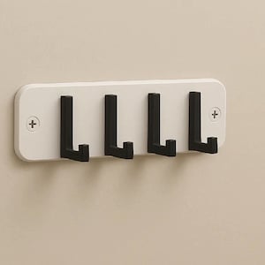 Puede incluir: Un perchero blanco montado en la pared con cuatro ganchos negros. El soporte rectangular con esquinas redondeadas se fija a la pared con tornillos. Los ganchos están espaciados uniformemente y sobresalen.