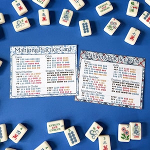Op de afbeelding: Een bovenaanzicht van ivoorkleurige Mahjong-tegels verspreid over een blauw oppervlak, met twee "Mahjong Practice Card" vellen. De tegels tonen verschillende symbolen en cijfers, sommige met bloemen en draken. De kaarten vermelden combinaties.