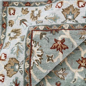 Könnte beinhalten: Nahaufnahme eines Teils eines hellblauen Teppichs mit floralem Muster. Der Teppich hat einen Rand mit einem sich wiederholenden Design in Braun-, Rot- und Grüntönen. Das florale Muster umfasst Blumen und Blätter in ähnlichen Farben.