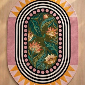 Könnte beinhalten: Ovaler Teppich mit floralem Muster. Der Teppich hat ein grünes Mittelfeld mit Blumen und Blättern, umgeben von einem rosa Rand, schwarz-weißen Streifen und gelben Dreiecken. Dieser Teppich ist für die Inneneinrichtung bestimmt.