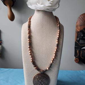 May include: A copper-colored necklace with a large, round pendant featuring an intricate design. The necklace is displayed on a mannequin with a white rose on top. The beads are a similar copper color, and the necklace has decorative spacers.
