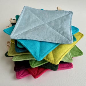 May include: A pile of square pot holders in shades of blue, green, and red. Each pot holder features a quilted diamond design and a small loop for hanging. The pot holders are made of fabric.