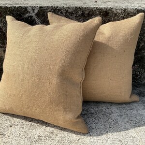 May include: Two square, tan burlap pillows are positioned on a concrete surface. The pillows have a textured appearance and are filled, with pointed corners. The pillows are a neutral color, suitable for various decor styles.