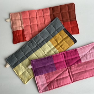 Puede incluir: Cuatro agarraderas rectangulares acolchadas en tonos de rojo, rosa, amarillo, gris y morado. Cada agarradera está dividida en secciones de bloques de color y tiene un pequeño lazo para colgar. Las costuras crean un patrón de cuadrícula.
