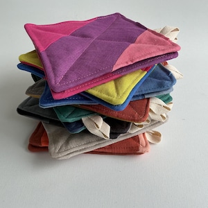 Puede incluir: Una pila de coloridos salvamanteles acolchados. Cada salvamanteles cuadrado presenta un patrón de diamantes y un lazo para colgar. Los colores incluyen rosa, morado, amarillo, azul, verde y naranja. Los salvamanteles son de tela.