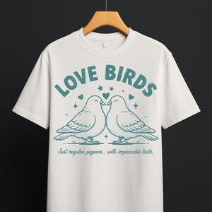 Könnte beinhalten: Ein cremefarbenes Kurzarm-T-Shirt mit einer blaugrünen Grafik von zwei Tauben, die sich gegenüberstehen. Der Text lautet oben LOVE BIRDS und unten Just regular pigeons... with impeccable taste., verziert mit kleinen Herzen und Sternen.