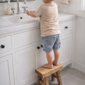 Könnte beinhalten: Ein Kind steht auf einem kleinen, rustikalen Holzschemel, um das Waschbecken in einem weißen Badezimmer zu erreichen. Das Kind trägt ein beiges T-Shirt und Jeansshorts. Eine Chromarmatur ist sichtbar.
