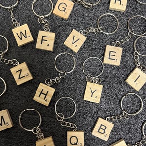 Könnte beinhalten: Schlüsselanhänger mit Holz-Scrabble-Steinen, die jeweils einen Buchstaben und einen Punktwert anzeigen. Die Steine sind mit silberfarbenen Ketten und Schlüsselringen verbunden. Sichtbare Buchstaben sind W, F, V, E, Y, B, P, Z, H, J, Q, G, R, I und M.