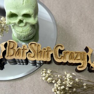Puede incluir: Un letrero de madera con la frase "Bat Shit Crazy" en un diseño tallado en capas. Dos siluetas de murciélagos flanquean el texto. Una calavera verde y flores secas están en el fondo, sobre una superficie reflectante.