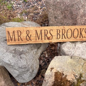 May include: A rectangular wooden sign with a natural light brown wood grain finish. The text MR & MRS BROOKS is engraved in a dark brown, serif font across the center of the board, which features a horizontal wood plank texture.