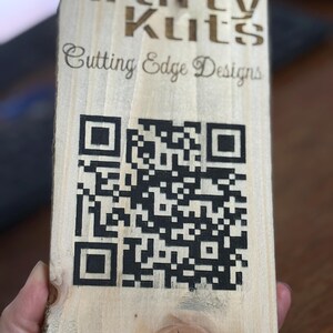Peut inclure: Panneau rectangulaire en bois clair avec le texte "Krafty Kuts Cutting Edge Designs" et un grand code QR noir. Le bois présente des nœuds naturels.