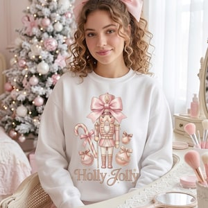 Peut inclure: Un sweat-shirt blanc à manches longues avec un motif de casse-noisette portant un grand nœud rose, entouré de sucres d'orge et de boules de Noël, avec le texte "Holly Jolly" imprimé en dessous dans des tons rose et doré.
